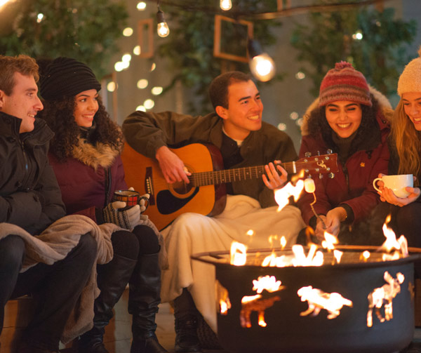 diverse group of people smiling, sitting around a fire with guitar
