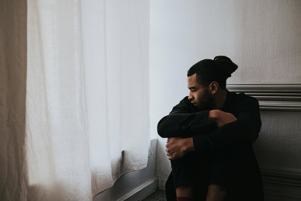 Man sitting on the floor by a window, looking pensive and reflective, symbolizing an emotional struggle