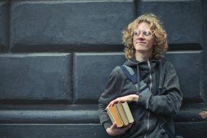 Young adult with curly blond hair and glasses, wearing a gray hoodie, holding a stack of books.