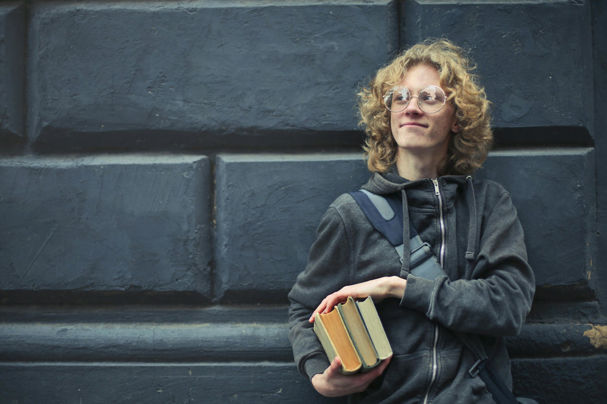 Young adult with curly blond hair and glasses, wearing a gray hoodie, holding a stack of books.