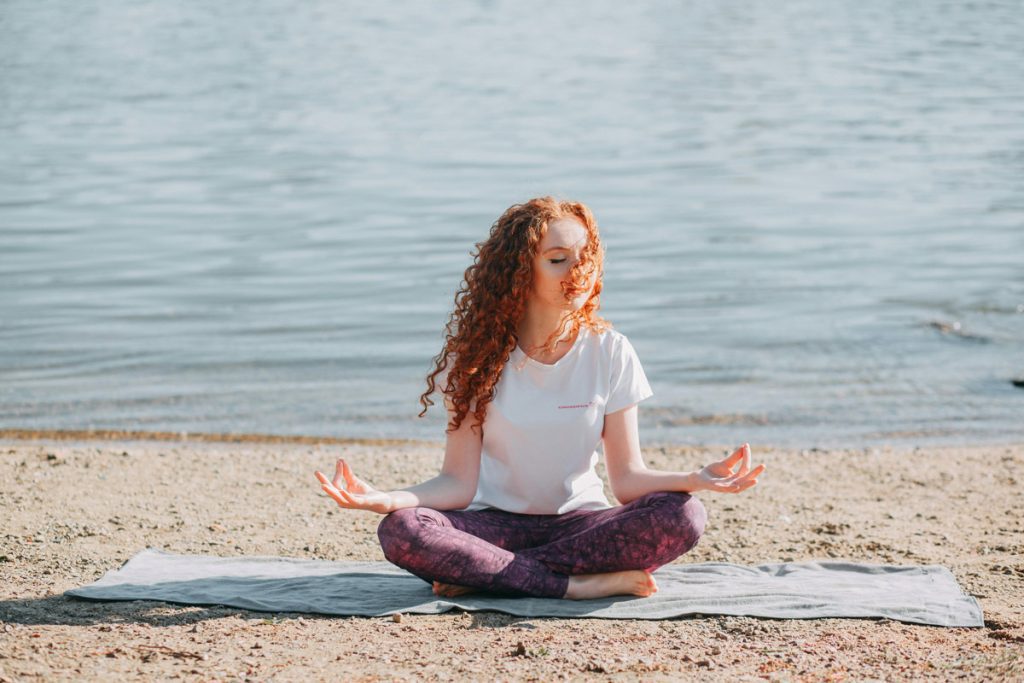 Young woman practicing trauma-aware yoga by the water, sitting cross-legged with eyes closed and hands resting on knees, finding calm and connection.