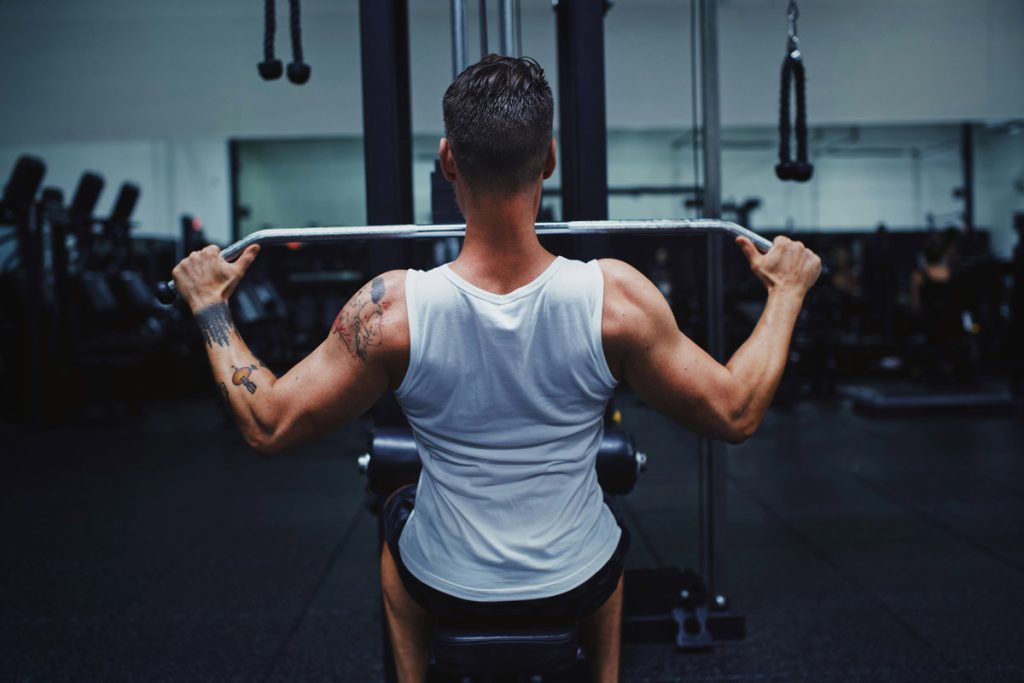 Man in a white tank top doing a lat pulldown exercise at the gym.
