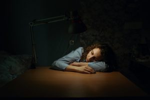 A young woman rests her head on a wooden desk, eyes closed, exhausted and emotionally vulnerable.