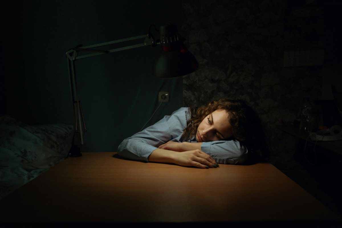 A young woman rests her head on a wooden desk, eyes closed, exhausted and emotionally vulnerable.