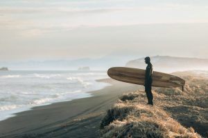 Man in contemplative pose holding surfboard on beach, representing mindfulness and healing journey from brokenness to wholeness.