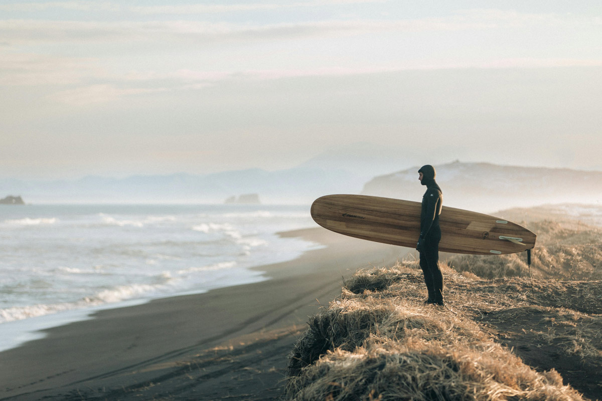 Man in contemplative pose holding surfboard on beach, representing mindfulness and healing journey from brokenness to wholeness.
