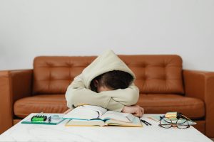 Student in beige hoodie with head down on desk surrounded by study materials including books, calculator, and glasses, depicting the exhaustion and overwhelm associated with executive dysfunction.