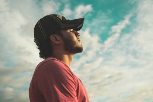 Man in red shirt and black cap practicing mindful breathing outdoors with eyes closed against a cloudy blue sky