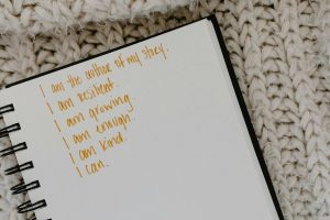 Person writing positive affirmations in a notebook on a cozy knitted blanket, representing daily mindfulness and self-care practices for emotional resilience.