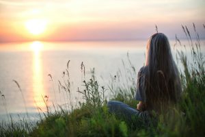 Person sitting peacefully in grass by water at sunset, practicing mindfulness during addiction recovery.