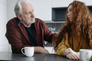 Older man with hand on shoulder of younger woman having supportive conversation at table.