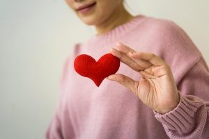 Person in pink sweater holding red heart shape over chest, representing heart health and self-care.