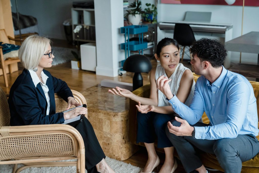 Couple sitting on couch during marriage counseling session with therapist in background.