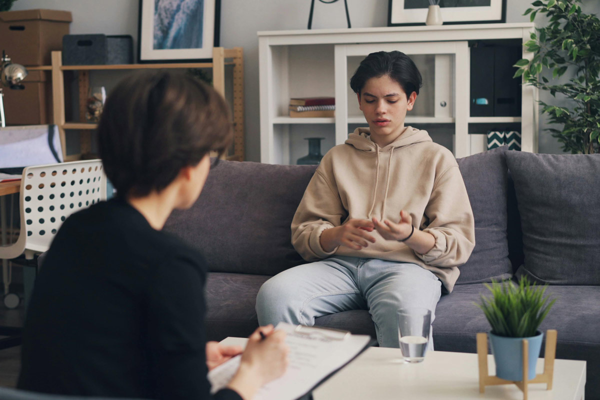 Teen in hoodie sitting on couch speaking with a psychiatrist.