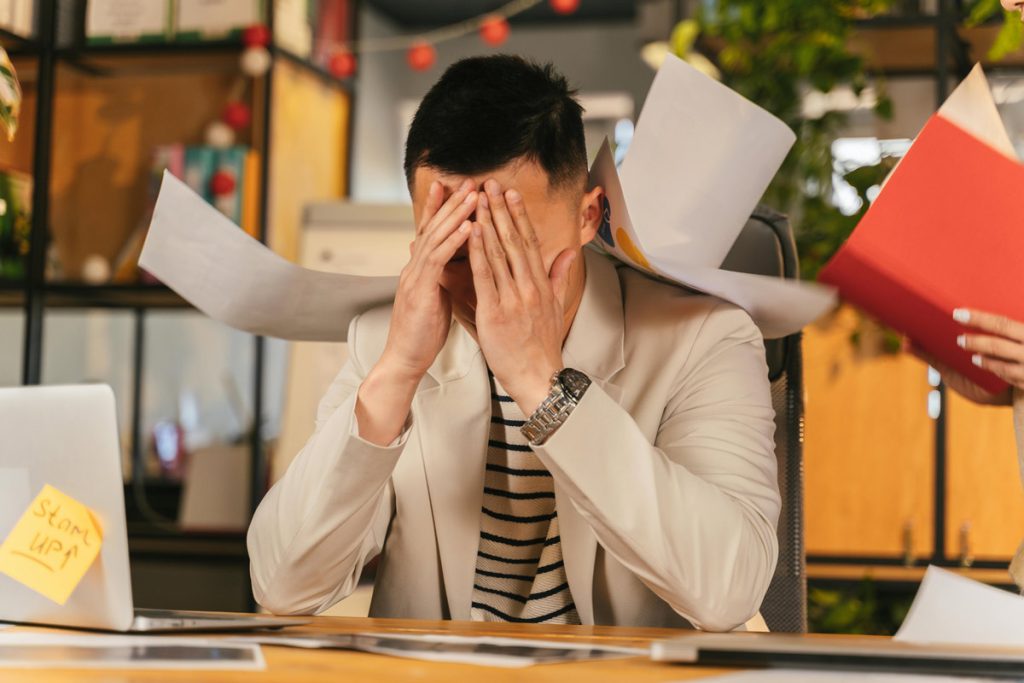 Stressed professional with head in hands at desk surrounded by scattered papers after making a mistake at work