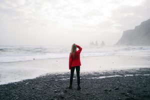 Person in red jacket standing on beach looking out at ocean and cliffs, representing reflection and the journey of changing unhealthy habits
