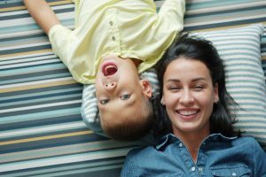 Mother and young child lying on striped blanket laughing together, demonstrating shared joy and emotional connection.
