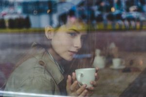 A young woman sitting in a café holds a coffee mug and gazes out through a rain-streaked window, her expression thoughtful and introspective, with blurred reflections visible in the glass.