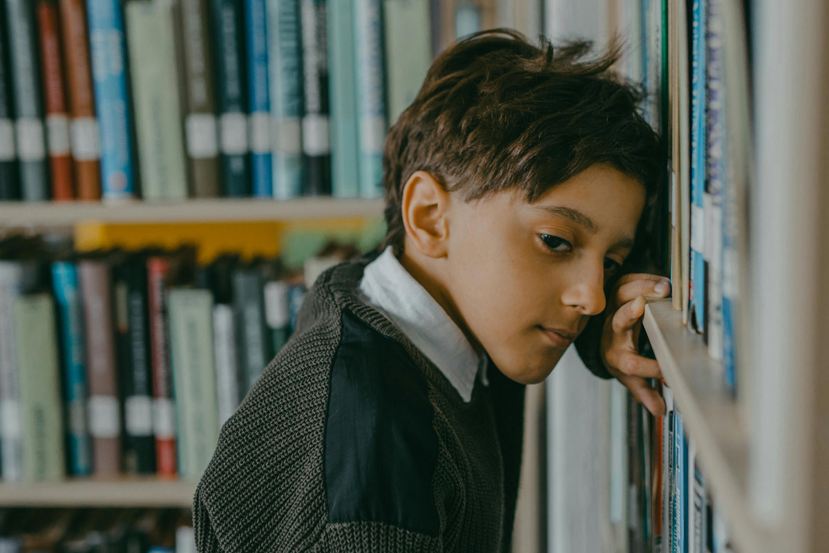 A young boy leans his head against a library bookshelf, looking sad and withdrawn.