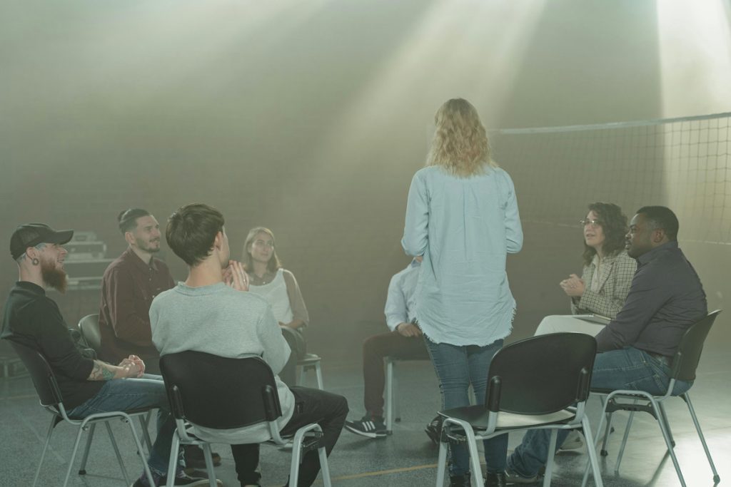 A diverse group of people seated in a circle for a community support group meeting, with one woman standing and addressing the group.