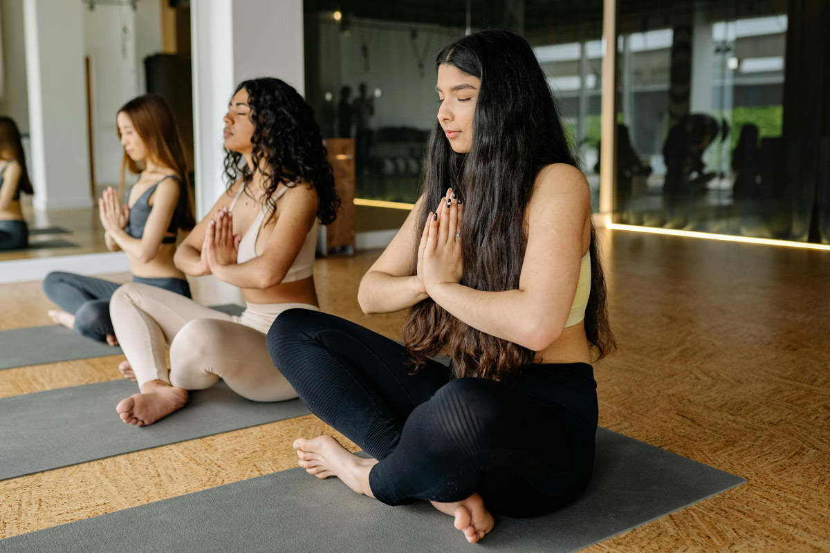 Three women sitting cross-legged on yoga mats with hands in prayer position, eyes closed in meditation