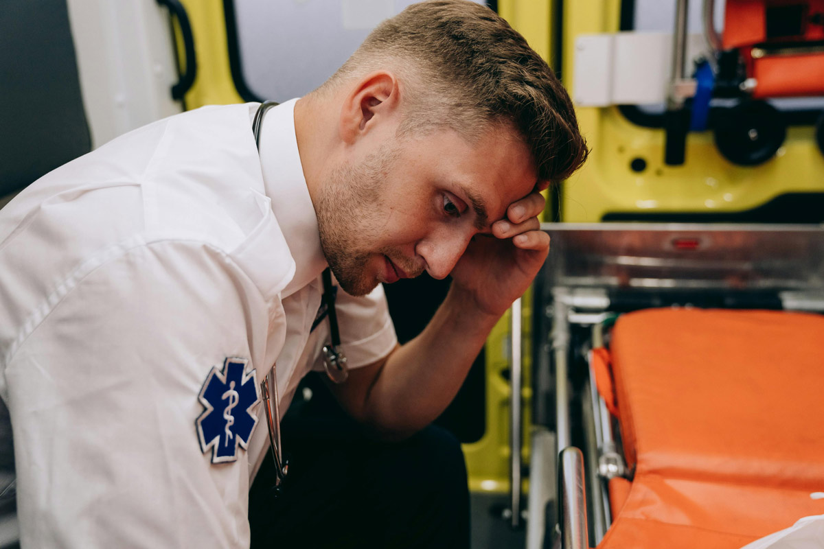 An EMT sits inside an ambulance with his head in his hand, reflecting the emotional burden of moral injury and PTSD among first responders.