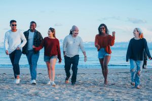 : A diverse group of six friends laughing and walking together on a beach, representing the social dimension of health in anxiety recovery.