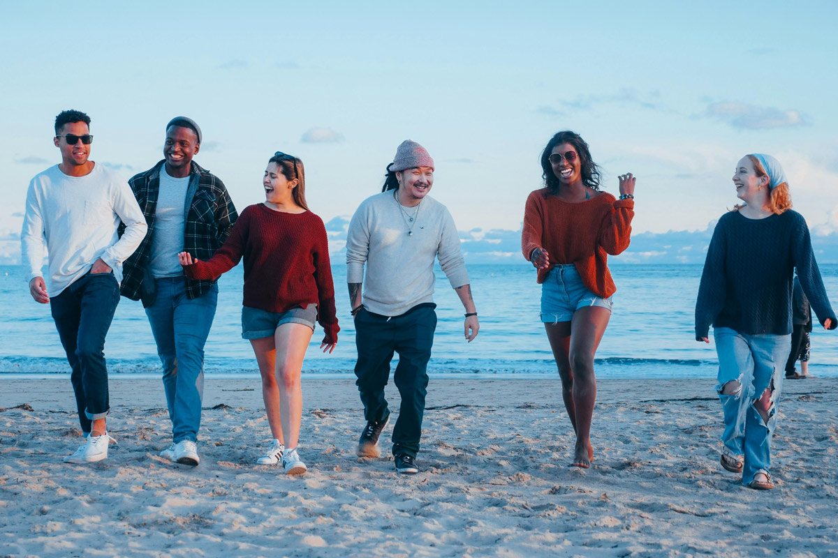: A diverse group of six friends laughing and walking together on a beach, representing the social dimension of health in anxiety recovery.