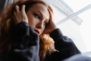 A young woman sits by a window, hands at her temples, looking down in quiet reflection — evoking the long-term emotional weight of childhood trauma.