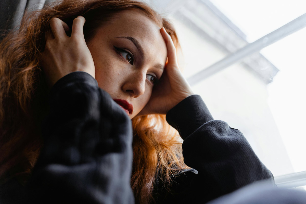 A young woman sits by a window, hands at her temples, looking down in quiet reflection — evoking the long-term emotional weight of childhood trauma.