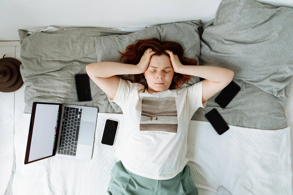 Young woman lying on a bed surrounded by laptops and smartphones, hands pressed to her head, eyes closed — overwhelmed by digital devices.