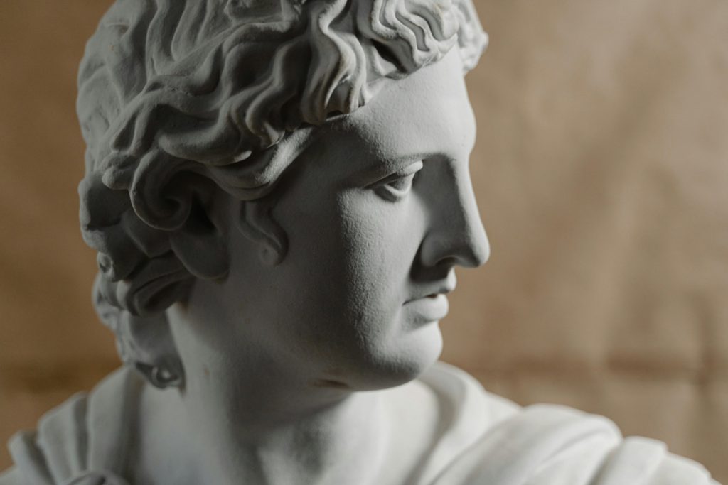 Profile view of a marble statue bust facing right with detailed curls and draped clothing on a warm background