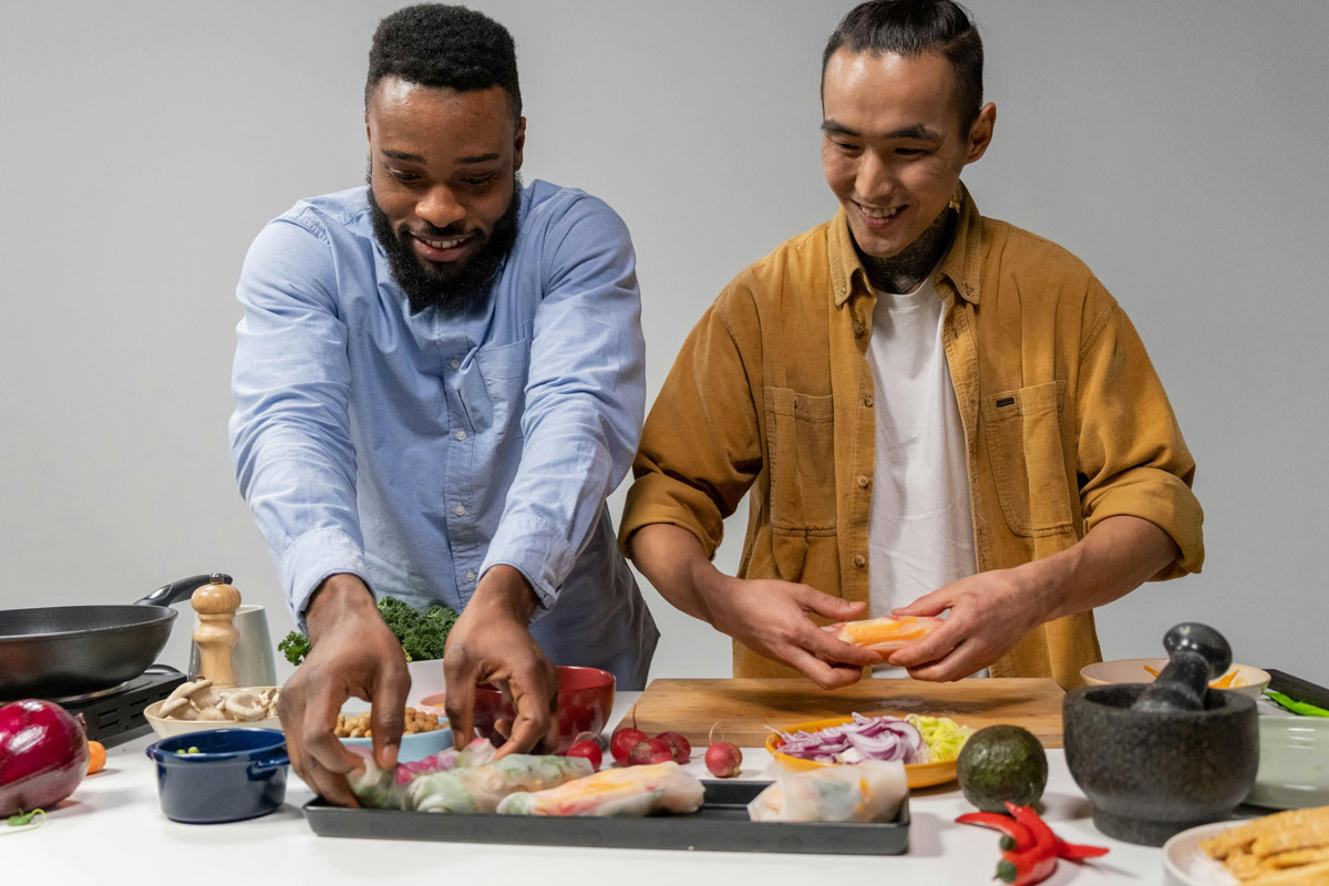 Two men stand at a kitchen counter smiling as they prepare fresh vegetables and wraps together, arranging ingredients on trays and cutting board.