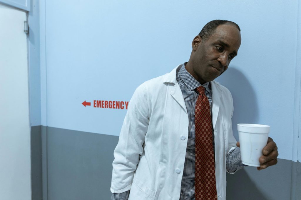 Man in a white lab coat with a red patterned tie stands in a blue hallway, holding a white disposable cup, with an 'EMERGENCY' sign and left-pointing arrow on the wall behind him.