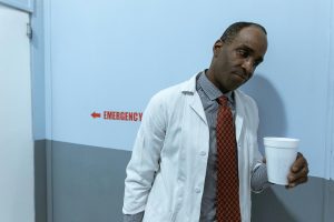 Man in a white lab coat with a red patterned tie stands in a blue hallway, holding a white disposable cup, with an 'EMERGENCY' sign and left-pointing arrow on the wall behind him.