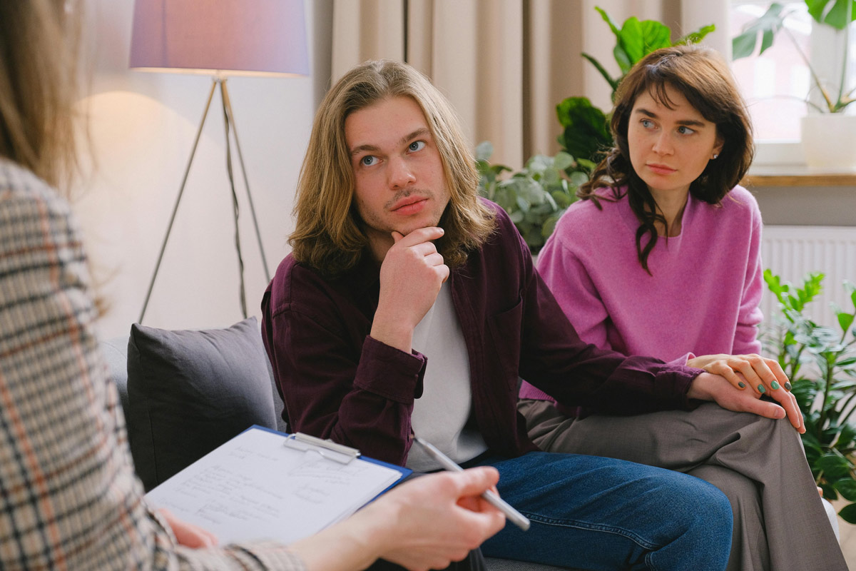 A man and woman sit on a couch across from a therapist taking notes during a therapy session.