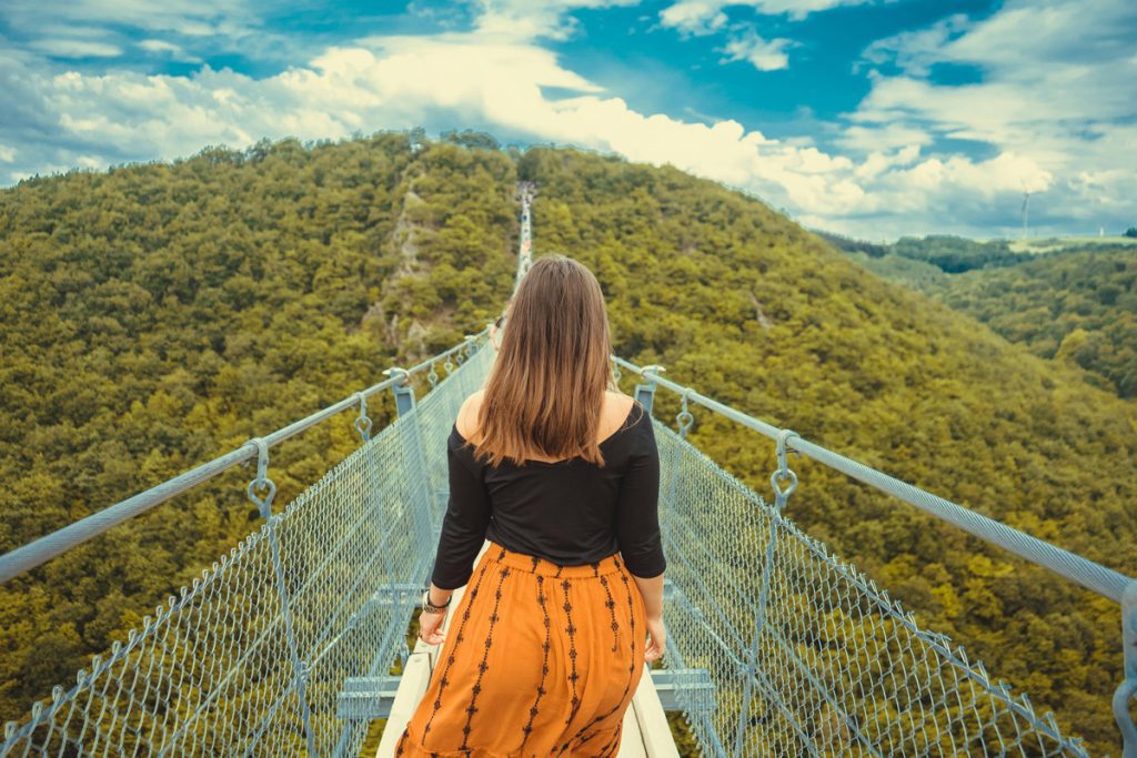 A young woman stands at the start of a long suspension bridge stretching toward a forested hillside, symbolizing the uncertain path of major life transitions in young adulthood.