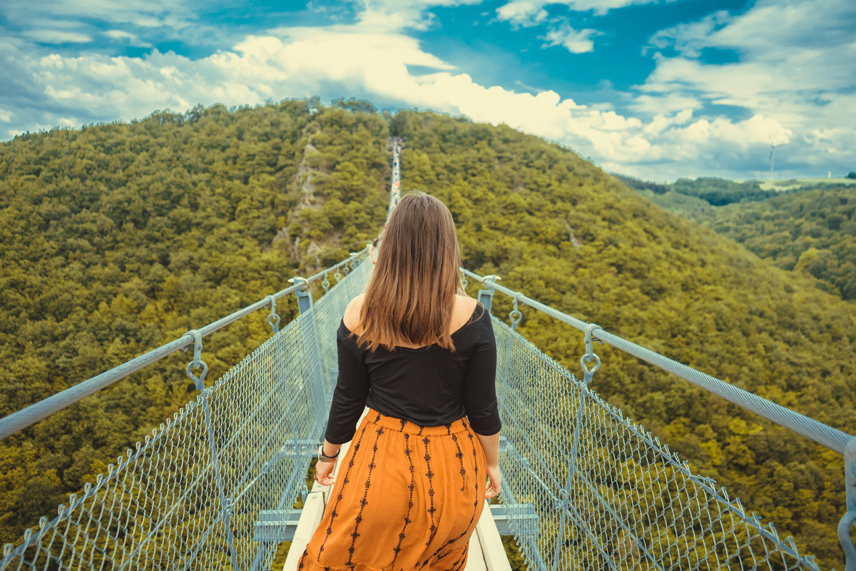 A young woman stands at the start of a long suspension bridge stretching toward a forested hillside, symbolizing the uncertain path of major life transitions in young adulthood.