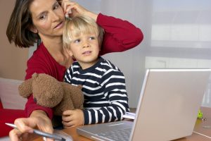 Mother adjusts an earpiece for her young son as they sit at a desk with a laptop, a stuffed bear, and a pen nearby.