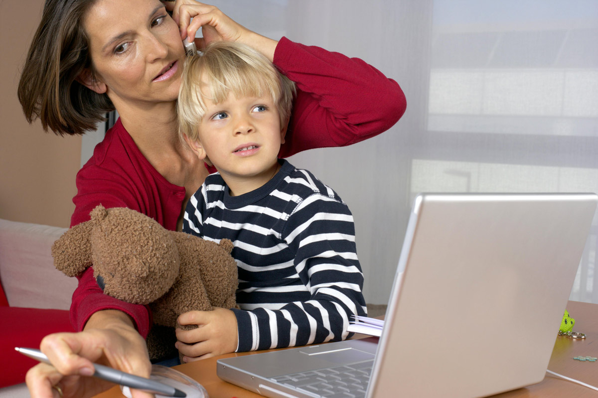 Mother adjusts an earpiece for her young son as they sit at a desk with a laptop, a stuffed bear, and a pen nearby.