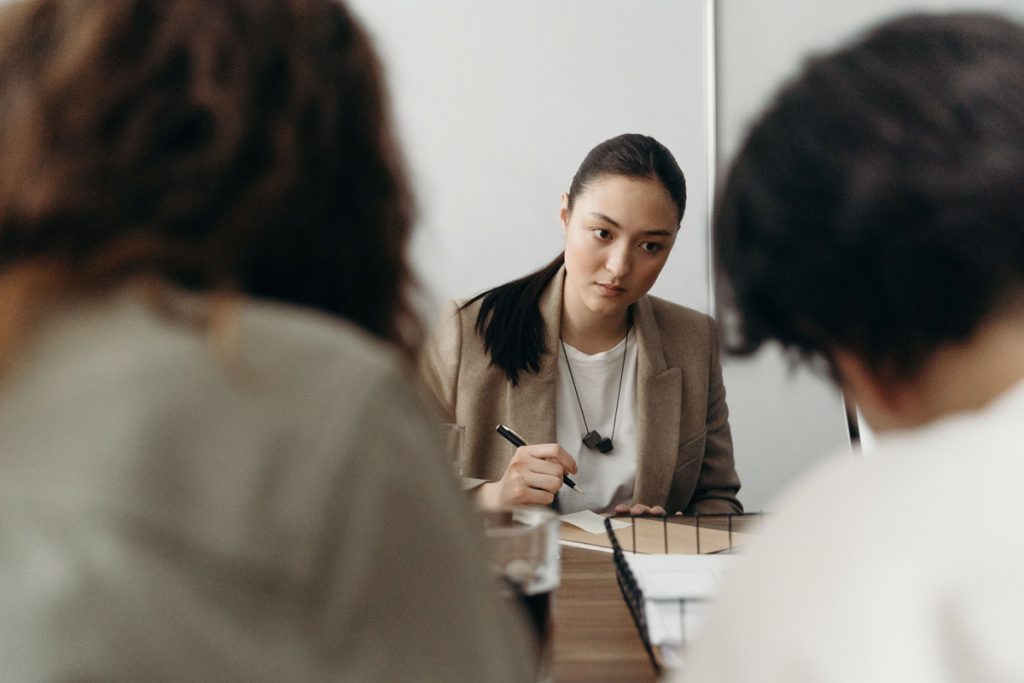 Woman in a blazer holding a pen, looking warily at blurred colleagues across the table, illustrating the self-doubt behind imposter syndrome.