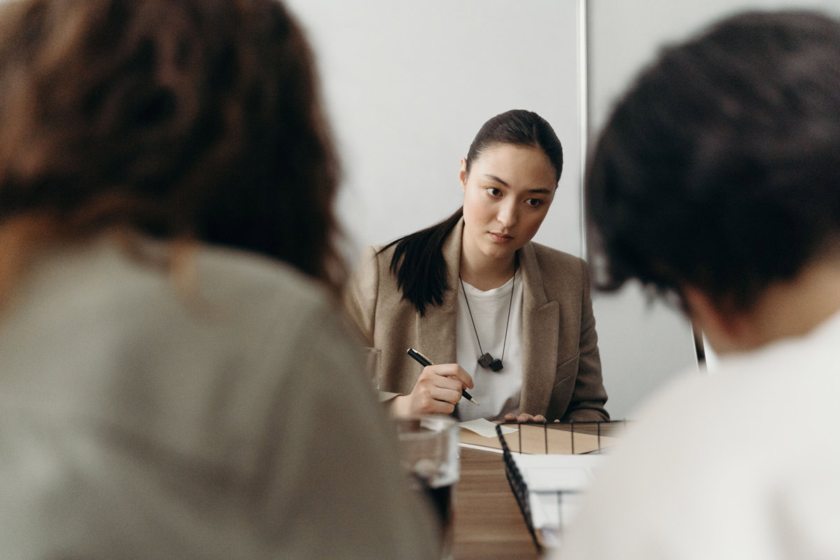 Woman in a blazer holding a pen, looking warily at blurred colleagues across the table, illustrating the self-doubt behind imposter syndrome.