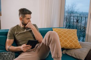 A man sits alone on a couch, holding a tablet and gazing out the window with a distant, preoccupied expression.