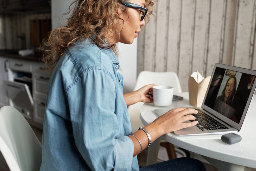 Woman with curly hair in a denim shirt video conferencing on a laptop, holding a mug at a white round table.