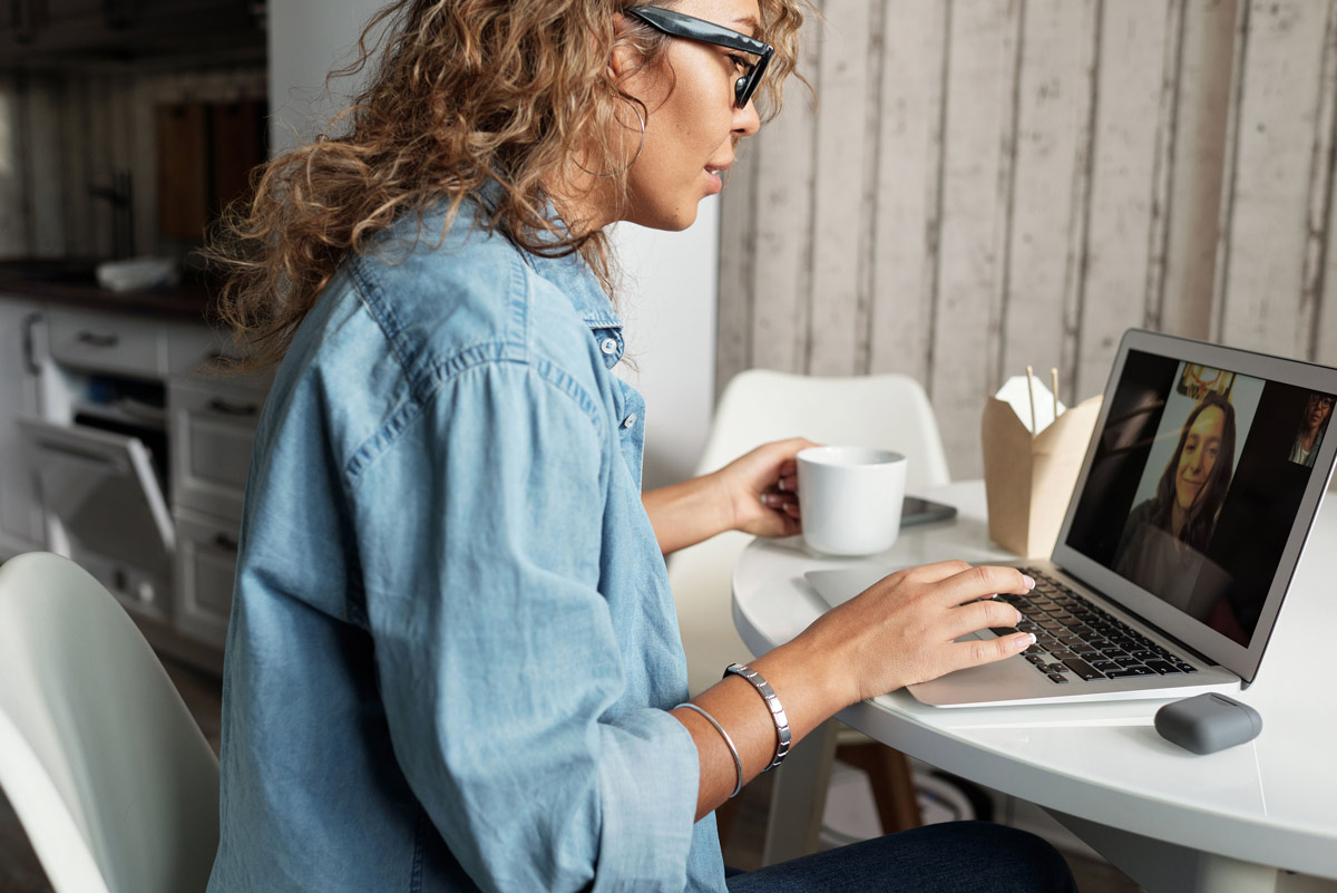 Woman with curly hair in a denim shirt video conferencing on a laptop, holding a mug at a white round table.