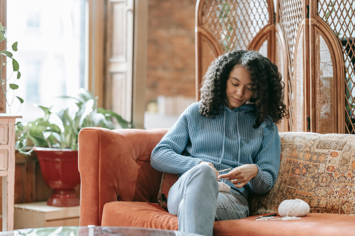 Young woman knitting on a couch at home, using the calming hobby as a mental health tool.