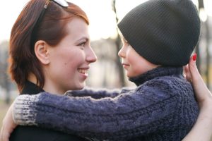 Woman and child share a warm hug outdoors, smiling at each other as the sun lights their faces (woman with red hair and sunglasses, child in a black beanie).”,