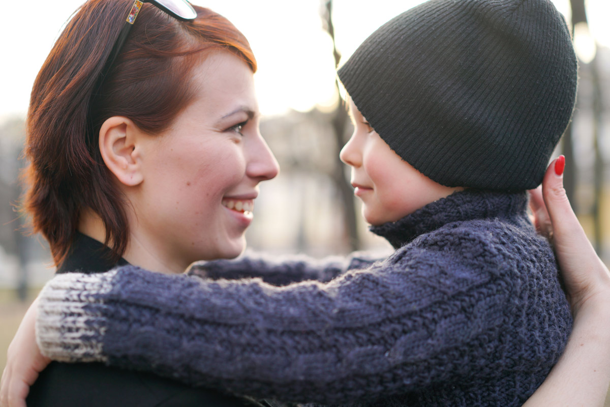 Woman and child share a warm hug outdoors, smiling at each other as the sun lights their faces (woman with red hair and sunglasses, child in a black beanie).”,