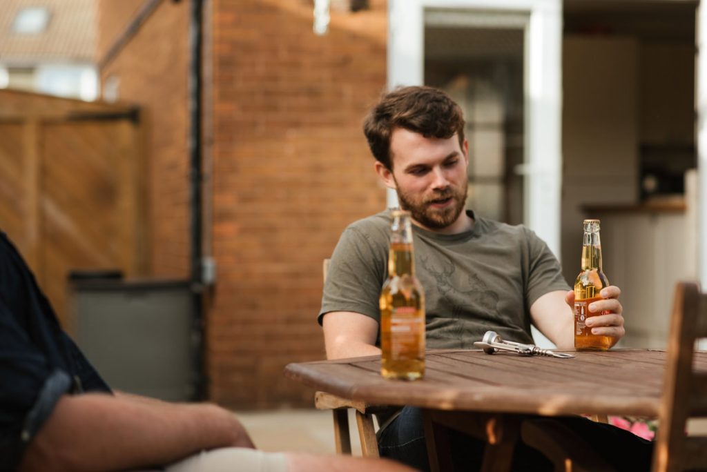 Young adult sitting outdoors at a table with a beer, talking with a friend