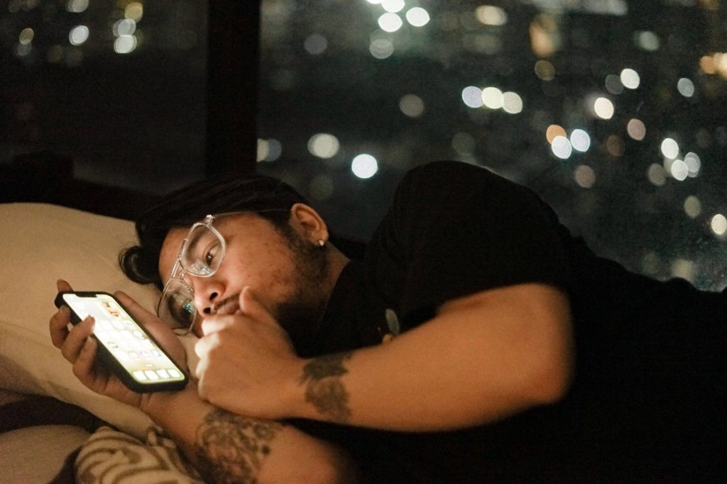 Man lying awake in bed at night, lit by his phone screen, city lights blurred in the window behind him.